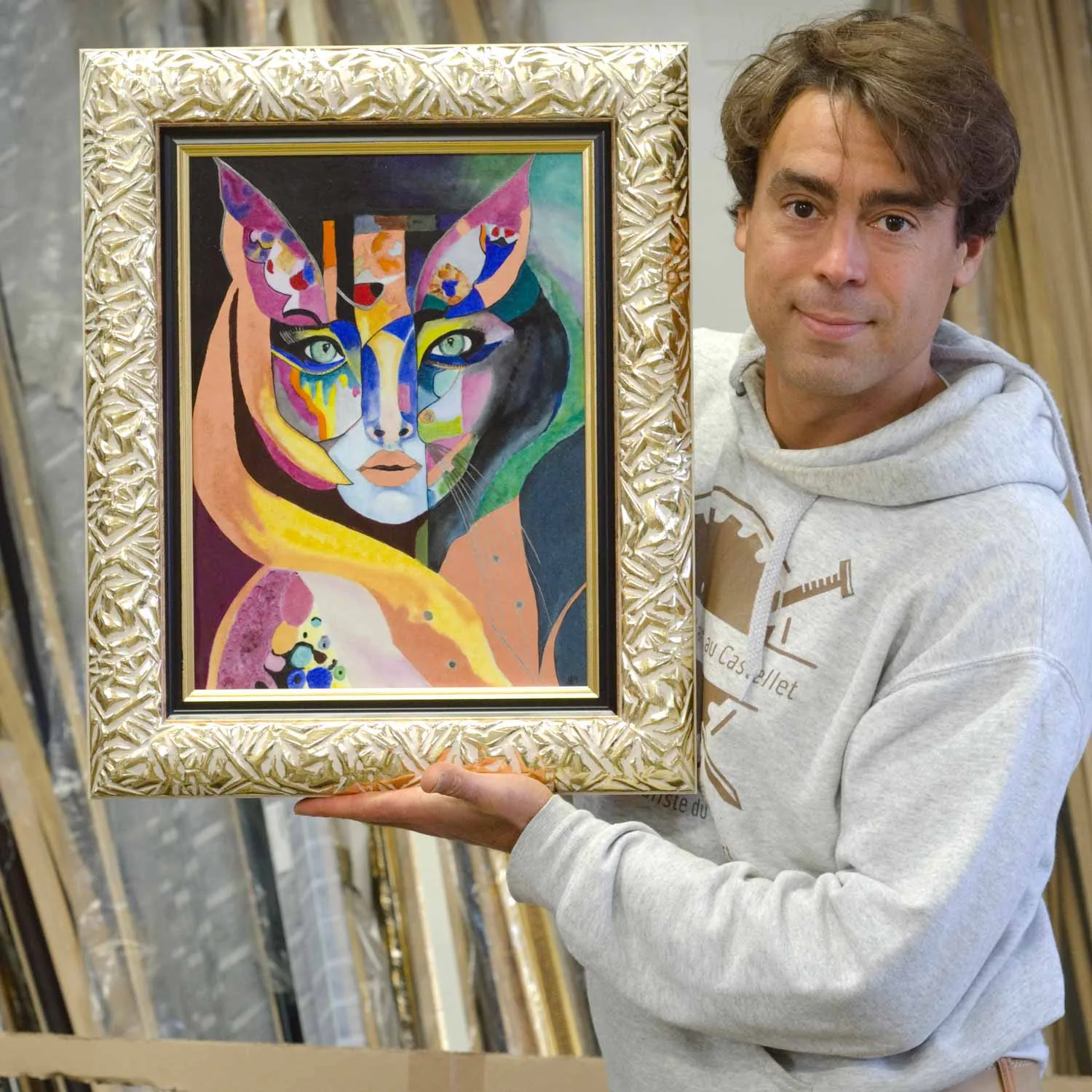 Julien Silvestro présente une toile colorée dans un cadre sculpté ivoire, devant les rayonnages de moulures de l'Atelier Valette